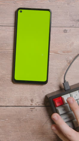 Vertical-Video-Overhead-Shot-Of-Table-And-Hands-Typing-On-Computer-Keyboard-With-Green-Screen-Mobile-Phone-